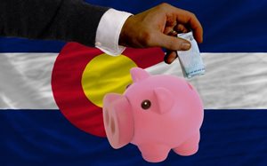 Hand putting money in a piggy bank in front of Colorado state flag